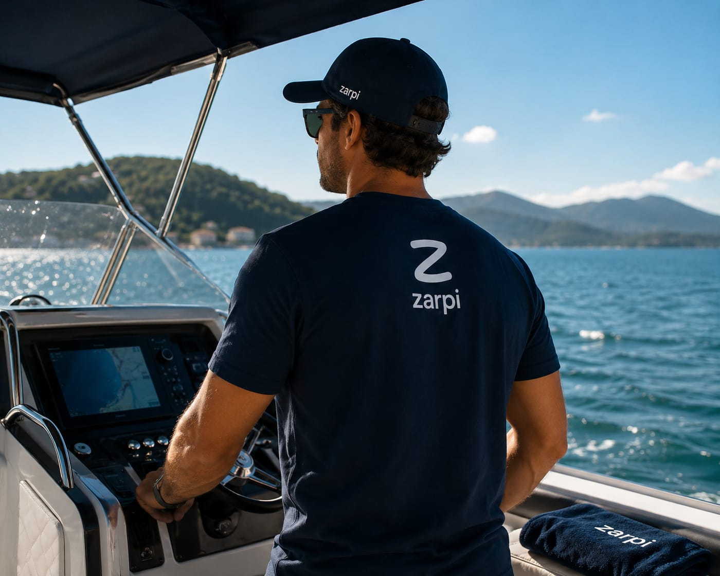 Marinheiro ao leme de uma lancha, de camiseta e boné da Zarpi, com a costa ao fundo